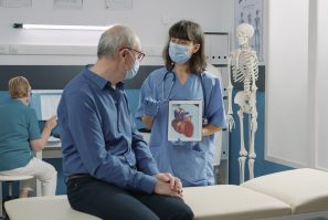 Female assistant holding tablet with cardiovascular system illustration, explaining heart condition to retired person during covid 19 pandemic. Nurse showing cardiology diagnosis on gadget.
